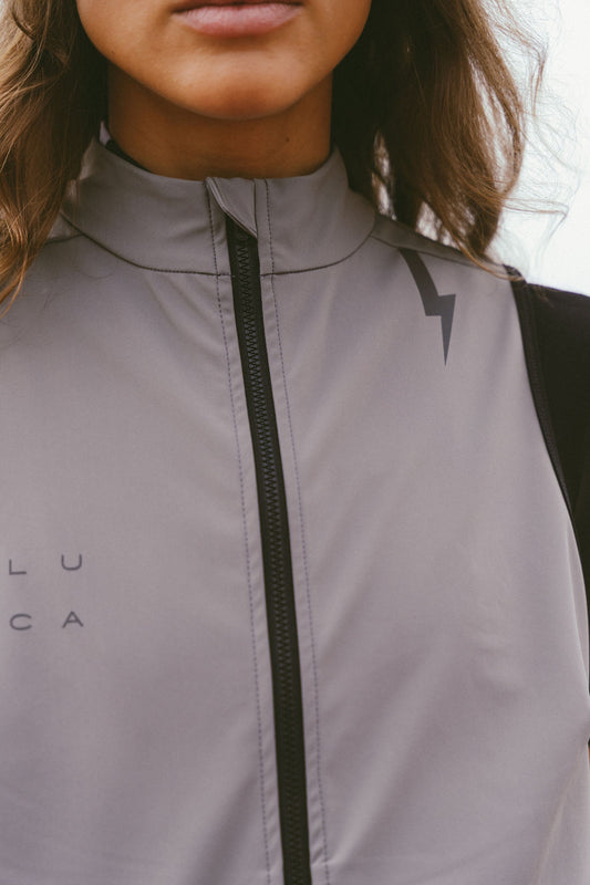 Close-up-detail-of-the-matte-taupe-grey-Luca-cycling-gilet-showcasing-the-full-length-black-zipper-and-high-collar-The-subtle-LUCA-brading-and-the-lighting-bolt-graphic-on-the-shoulder-are-visible-against-the-technical-fabric