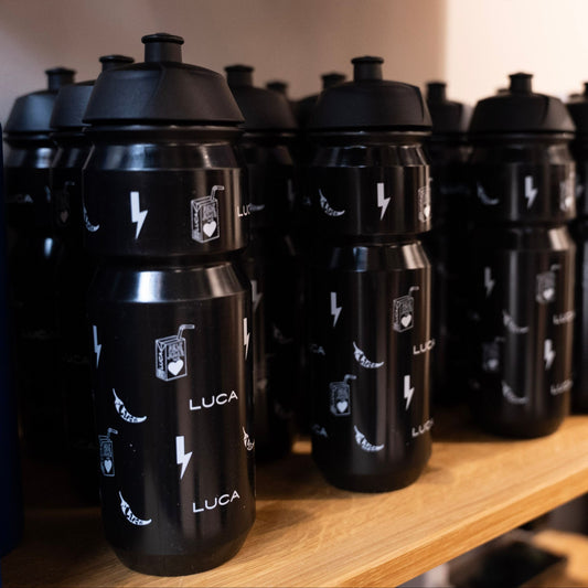 A-close-up-slightly-darker-photo-of- multiple-matte-black-cycling-water- bottles-displayed-on-a-light-wooden- shelf-The-bottles-are-branded-with-the-LUCA-logo-and-feature-playful- white-graphics-including-lightning- bolts-hearts-in-a-juice-box-design-and-a-chili- pepper-showcasing-the-unique- branding-pattern-in-a-bulk-setting