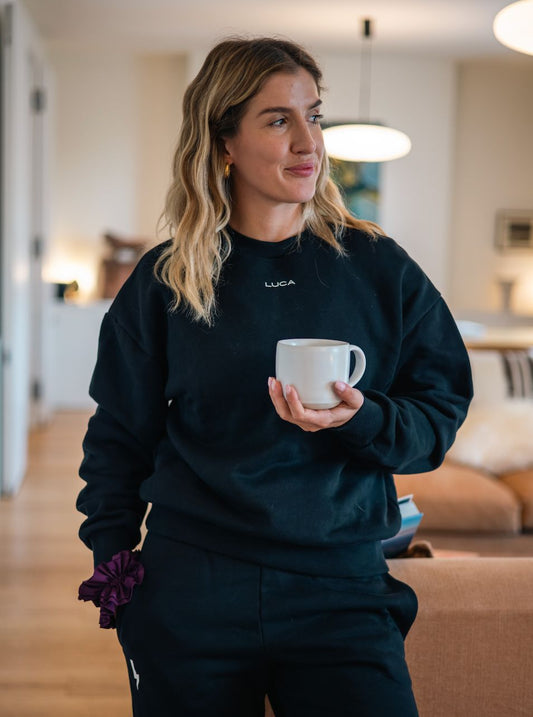 Woman-wearing-a-black-Luca-logo- sweatshirt-and-matching-sweatpants-holding-a-mug-indoors