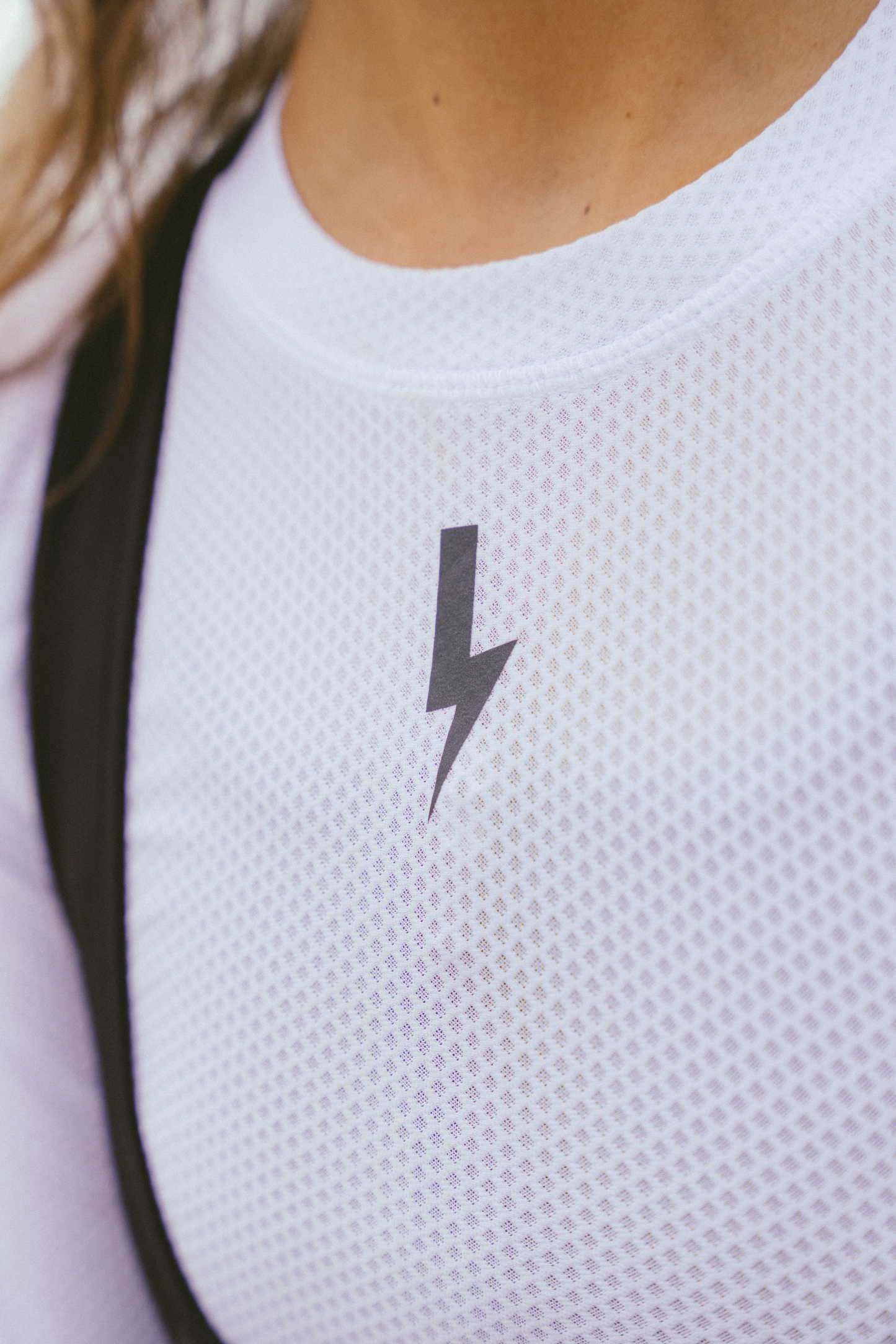 Extreme-close-up-of-a-white-Radiance-base-layer-top-showcasing-the-detailed-open-mesh-of-the-moisture-wicking- fabric-Centered-just-below-the-crew-neck-collar-is-a-prominent- black-lightning-bolt-sign-A-black- strap-likely-from-bib-shorts-is-visible-on-the-left-shoulder