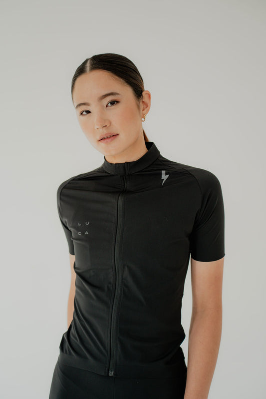 Young-woman-modeling-a-black-short-sleeve-cycling-jersey-with-a-full-zip-a-high-collar-and-a-small-lighting-bolt-sign-on-the-chest