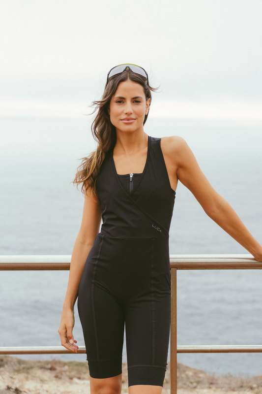 A-smiling-woman-models-a- sleeveless-black-cycling-bib-short- unitard-with-a-front-zipper-and- criss-cross-bodice-detail-standing- outdoors-by-a-coastal-railing- overlooking-the-ocean-She-is- wearing-mirrored-cycling-sunglasses-pushed-up-onto-her- head