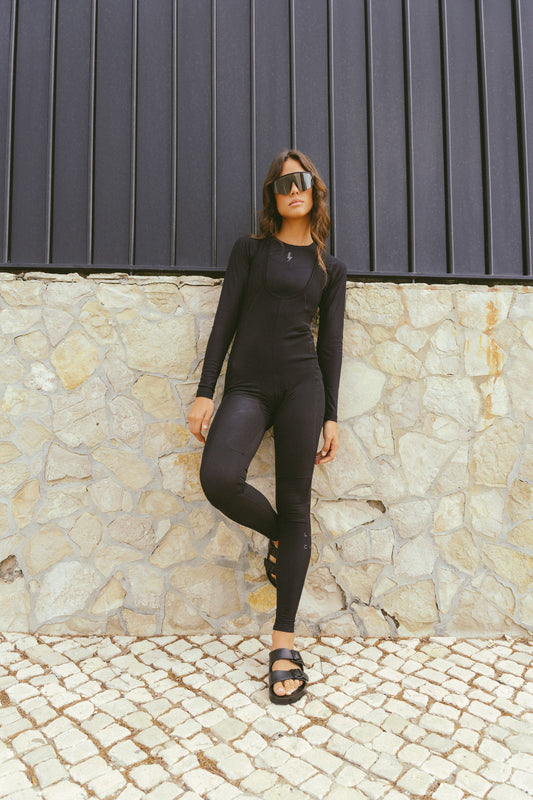 A-full-length-outdoor-lifestyle-photo-featuring-a-young-woman-with-wavy-brown-hair-wearing-a-fitted-long-sleeve-black-jumpsuit- unitard-with-full-length-legs-The- garment-appears-to-have-a-subtle- ribbed-textured-pattern-and-features-a-slightly-scooped-neck- with-layered-detailing-She-is- wearing-oversized-dark-sunglasses- and-black-two-strap-slip-on-sandals-(like-Birkenstocks)-She-is- leaning-against-a-textured-stone- wall-with-a-dark-slatted-fence-above-it-standing-on-a-cobblestone-pavement