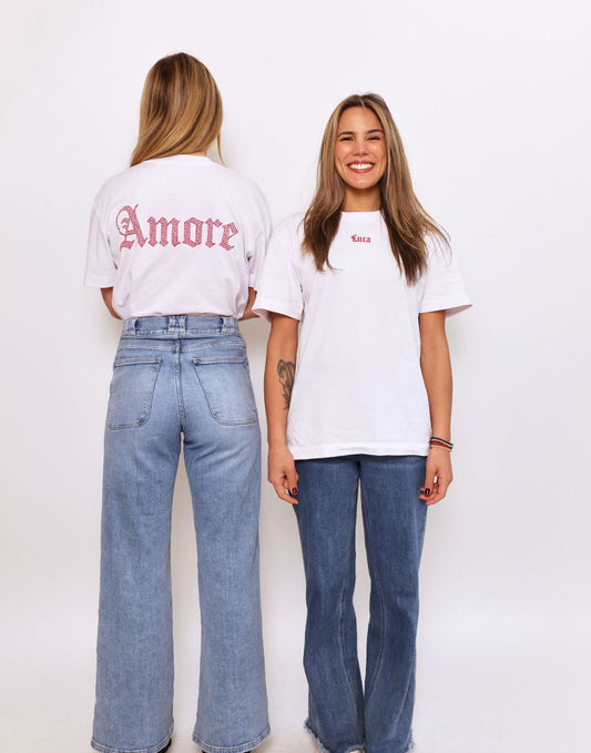 Two-women-modeling-white-graphic-t-shirts-paired-with-high-waisted-denim-jeans-One-t-shirt- shows-the-'Amore'-design-in-pink- gothic-text-on-the-back-the-other- shows-a-subtle-'Luca'-logo-on-the- front-chest