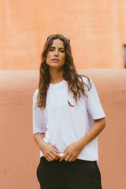 A-woman-with-long-brown-hair-stands-facing-forward-wearing-a- relaxed-fit-white-crewneck-t-shirt- featuring-a-small-vibrant-chili-pepper- embroidery-on-the-chest-She-is-also- wearing-black-pants-and-sunglasses- pushed-up-on-her- head-posing-against-a-muted- orange-wall