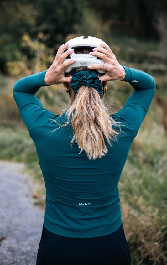 Back-view-of-a-cyclist-wearing-a-teal- long-sleeve-cycling-jersey-adjusting-a-white-helmet-showing- the-LUCA-brand-logo-on-the back