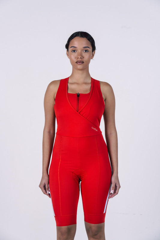 A-studio-shot-of-a-woman-modeling-a-bright-red-women's-cycling-all-in-one-against-a-plain-white-background-The-suit-features-a-sleeveless-top-with-a-racerback-design-a-short-front-zipper-and-a-unique-crossover-V-neck-detail-on-the-chest-for-support-The-integrated-shorts-have-a-mid-thigh-length-and-a-white-reflective-stripe-detail-on-the-outer-sides-of-the-legs