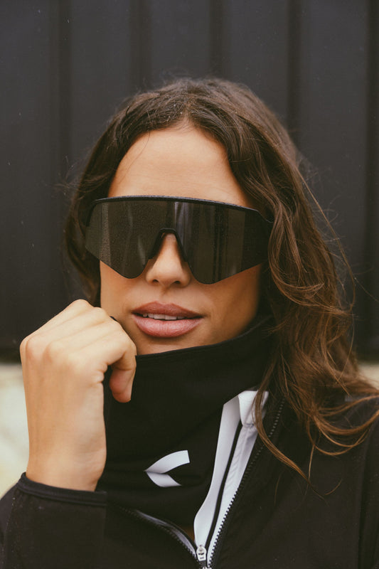A-close-up-portrait-of-a-woman-wearing-a-black-athletic-neck-gaiter-pulled-up-over-her-chin-and-large-black-shield-style-sport-sunglasses-She-holds-the-gaiter-near-her-neck-highlighting-its-snug-fit-and-versatile-design-for-cold-weather-The-black-gaiter-has-a-white-chevron-visible-near-the-chest-area-of-her-black-jacket