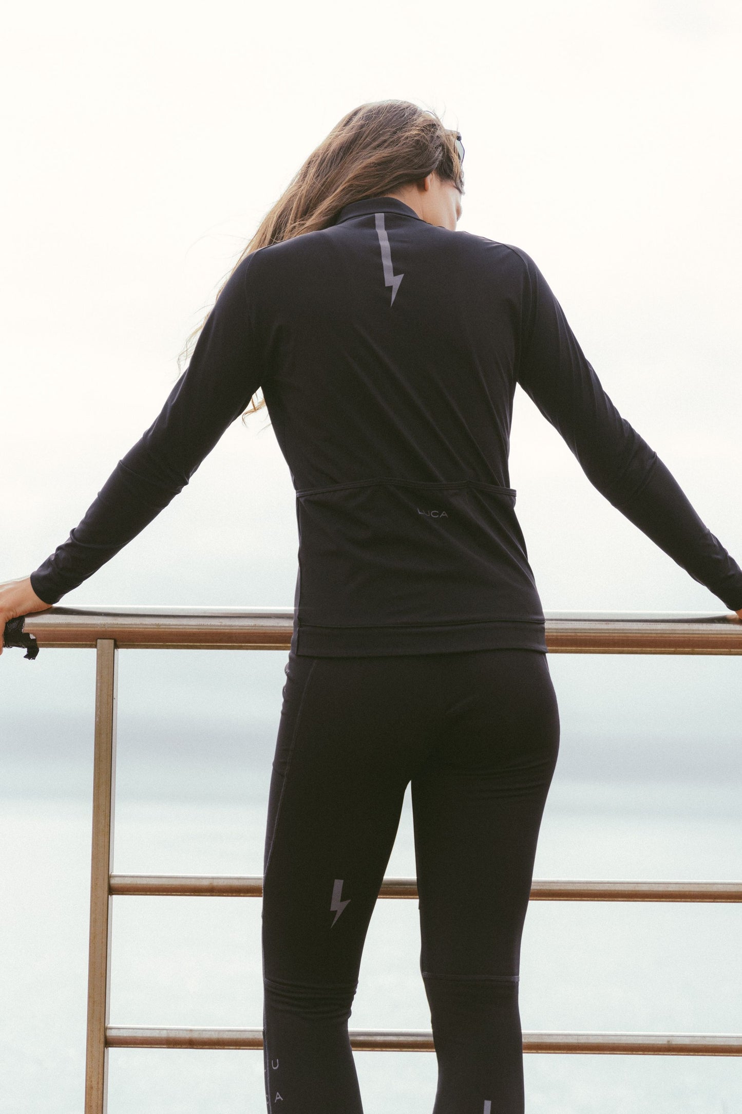 Full-back-view-of-the-model-wearing-the-black-Horizon-jersey-and-matching-leggings-showcasing-the-complete-outfit-the-image-highlights-the-reflective-lighting-bolt-sign-on-the-back-of-the-jersey-and-the-claves-of-the-pants-for-enhanced