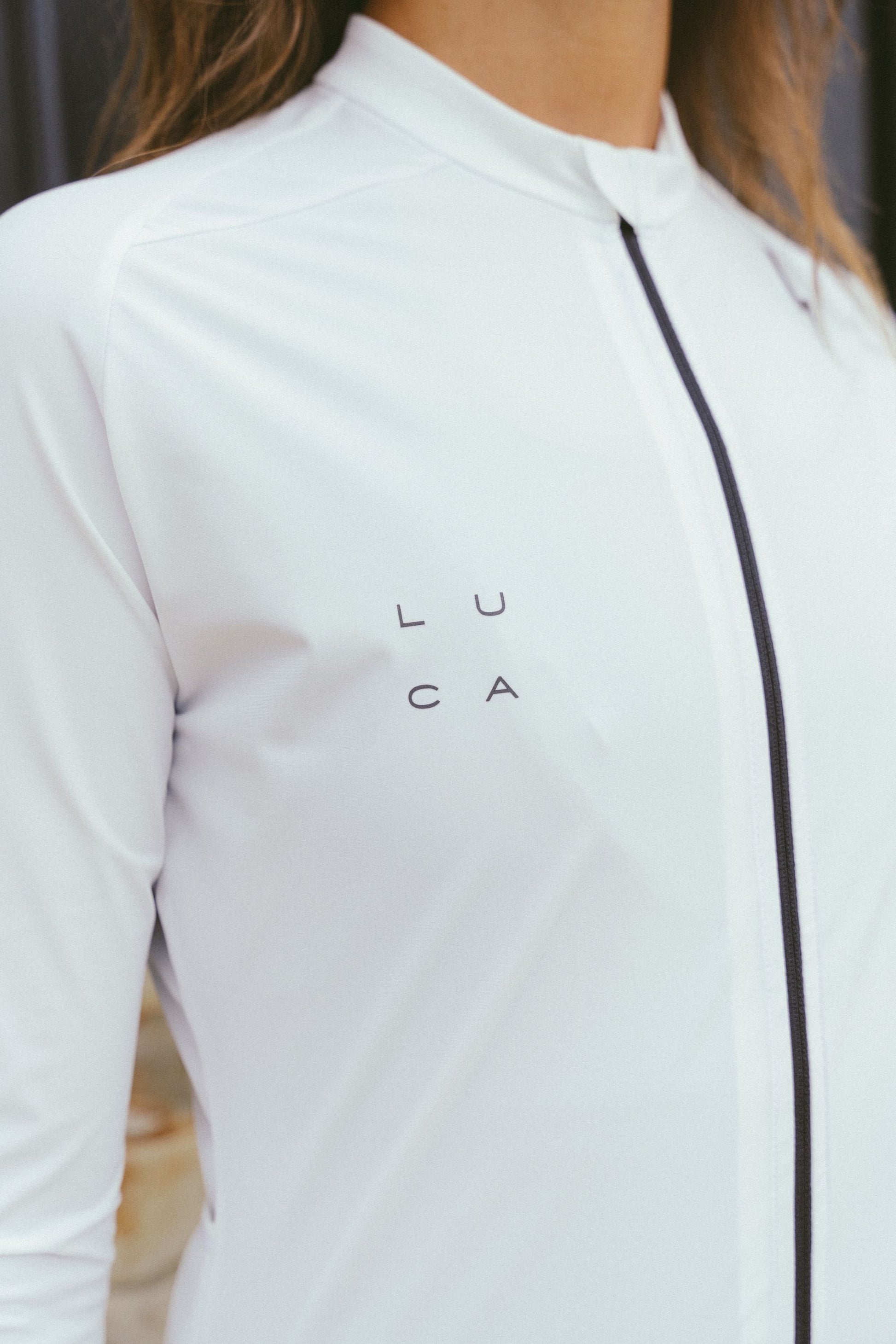 Close-up-detail-of-the-smooth-white-fabric-of-the-Luca-long-sleeve-cycling-jersey-showing-the-black-full-length-zipper-and-the-square-arranged-LUCA-logo-printed-on-the-chest