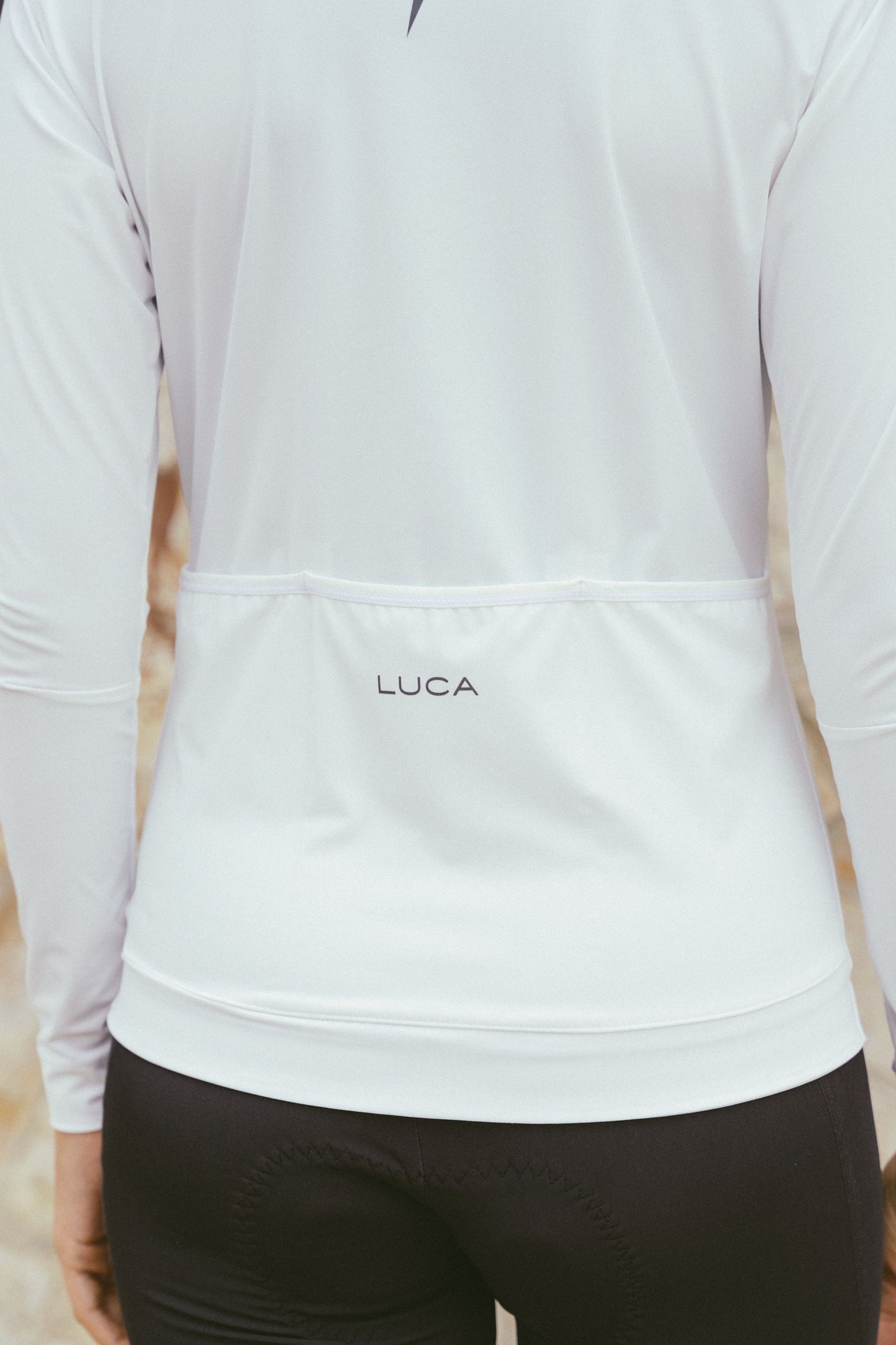 Close-up-of-the-lower-back-of-the-white-Luca-long-sleeve-cycling-jersey-clearly-showing-the-elasticated-rear-pocket-line-the-subtle-LUCA-branding-prited-above-the-gripped-hem-to-keep-the-jersey-in-place-during-a-ride
