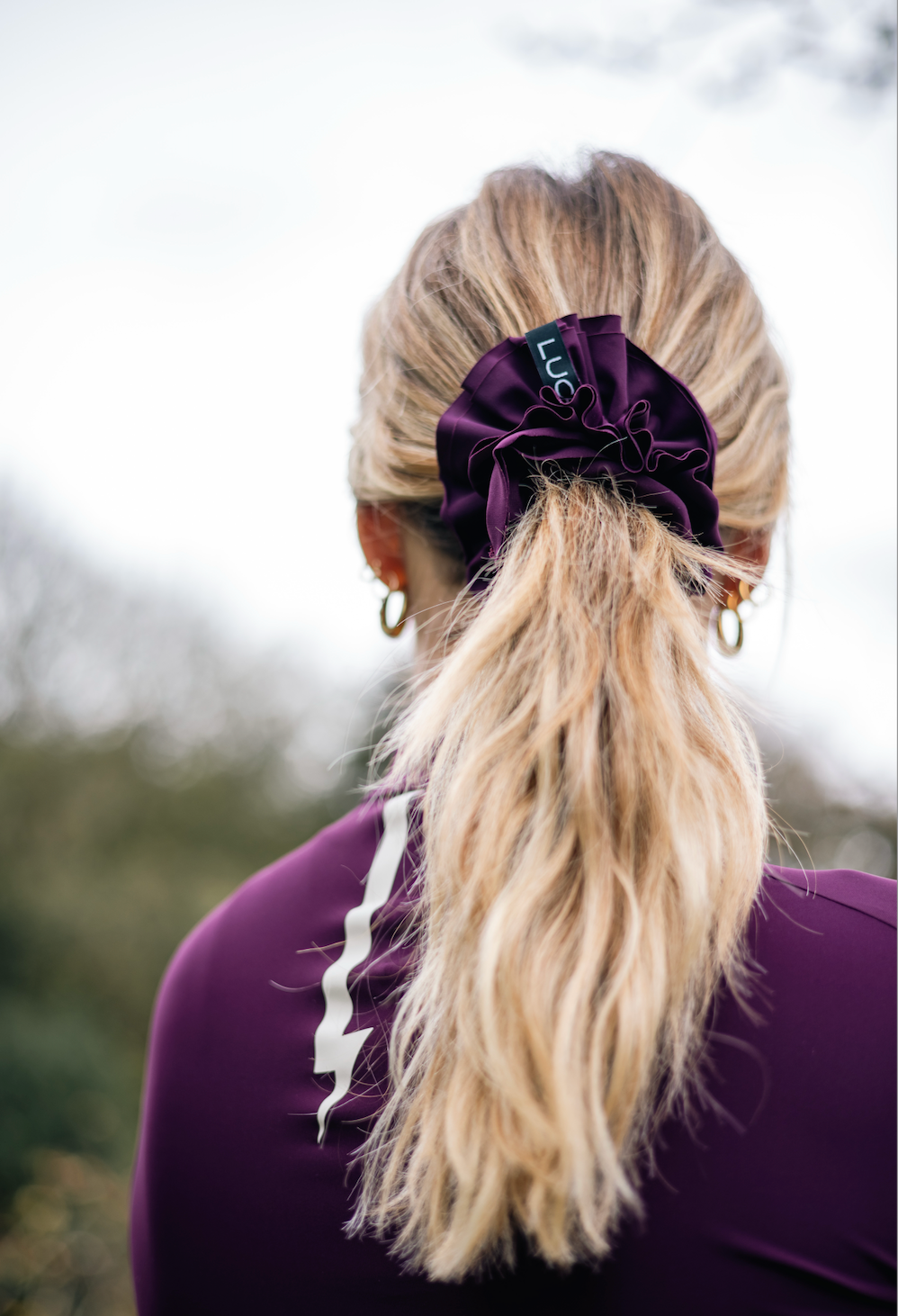 Woman-wearing-a-purple-athletic- top-and-a-large-dark-purple-Luca- logo-Uplift-scrunchie-in-her-ponytail