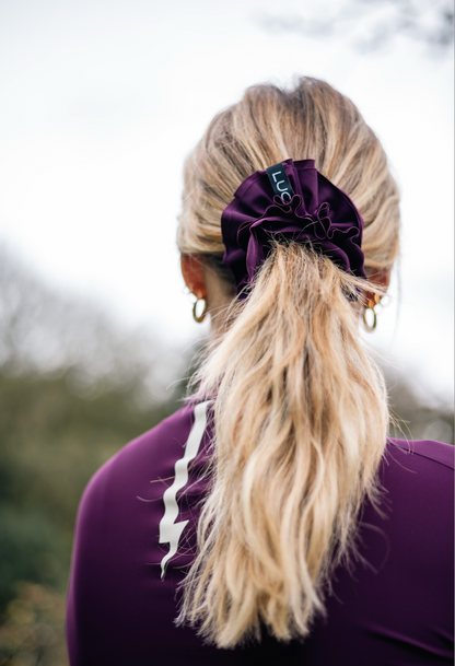 Woman-wearing-a-purple-athletic- top-and-a-large-dark-purple-Luca- logo-Uplift-scrunchie-in-her-ponytail