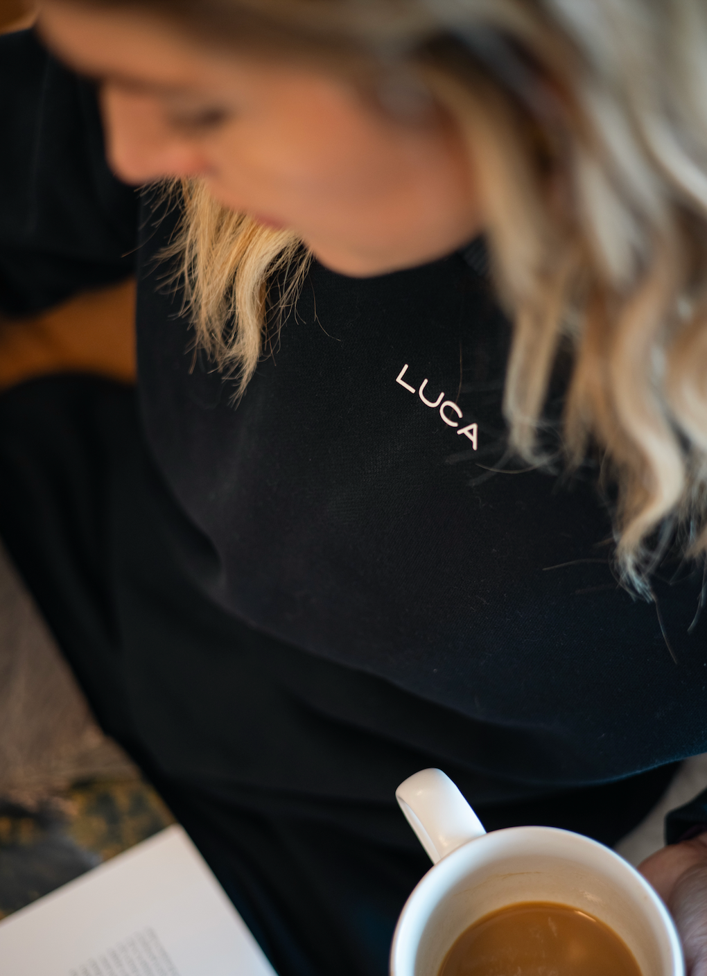 Close-up-of-a-woman-wearing-a- black-sweatshirt-with-the-Luca-logo-embroidery-while-holding-a- coffee-cup