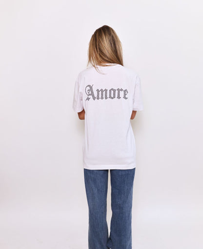 White-t-shirt-with-"Amore"-written- across-the-back-in-a-sparkling-gothic-style-rhinestone-diamond-look-font-shown-on-a-person- wearing-blue-jeans