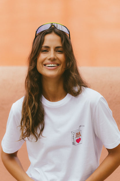 A-beautiful-image-of-a-smiling-woman-wearing-a-white-crew-neck-t-shirt-with-a-SELF-LOVE-JUICE-graphic-print-on-the-chest-The-print-on-the-chest-The-print-features-a-cute-retro-style-juice-box-with-a-red-heart-She-has-iridescent-mirrored-sports-sunglasses-pushed-up-on-her-head-and-stands-against-a-terracotta-