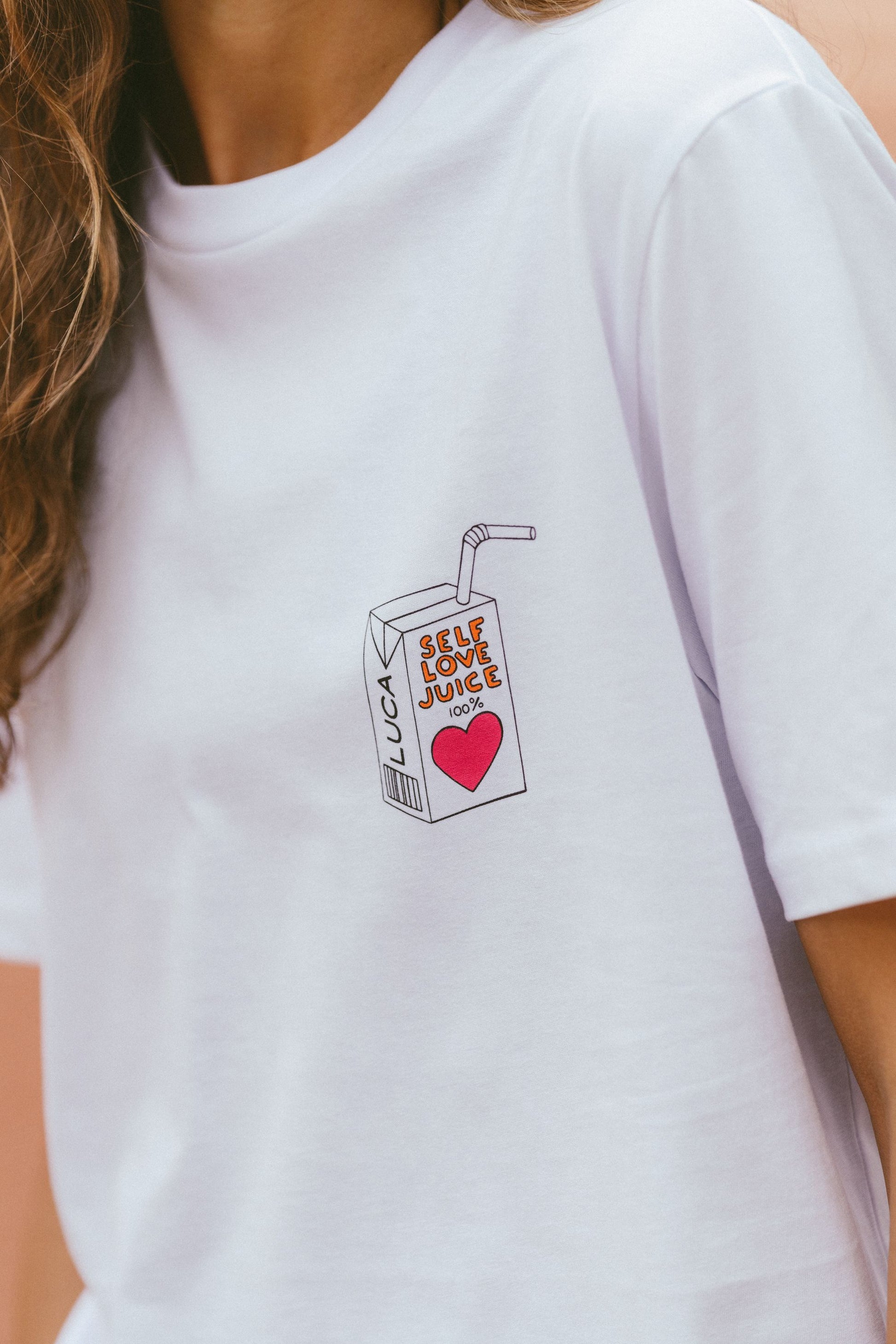 A-close-up-studio-shot-of-a-white-cotton-crew-neck-t-shirt-with-a-cetered-chest-graphic-The-detailed-graphic-depicts-a-retro-style-juice-box-with-a-straw-featuring-the-text-SELF-LOVE-JUICE-and-a-large-red--heart-The-brand-name-LUCA-is-printed-vertically-on-the-side-of-the-box-