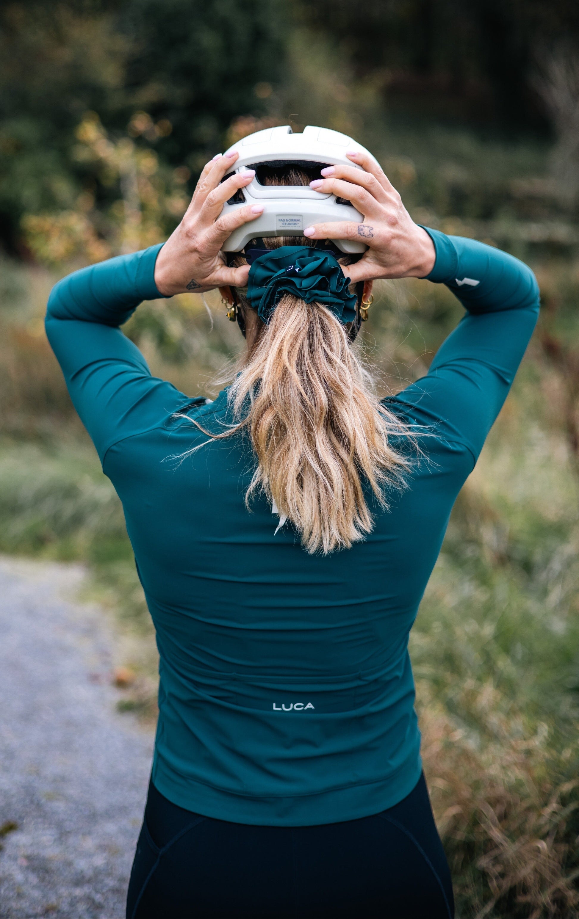Back-view-of-a-cyclist-wearing-a-teal- long-sleeve-cycling-jersey-adjusting-a-white-helmet-showing- the-LUCA-brand-logo-on-the back