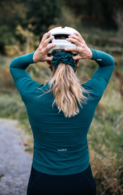 Back-view-of-a-cyclist-wearing-a-teal- long-sleeve-cycling-jersey-adjusting-a-white-helmet-showing- the-LUCA-brand-logo-on-the back