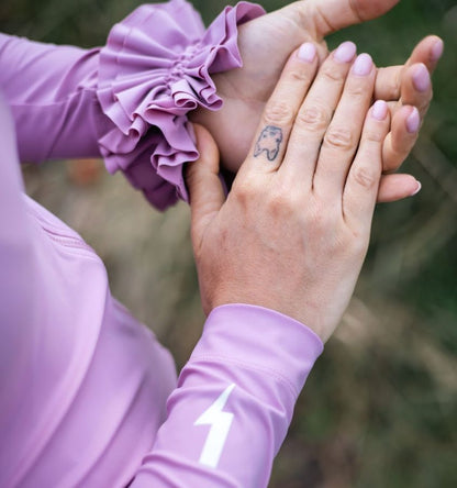 Close-up-of-a-hand-resting-on-a- light-purple-long-sleeve-cycling-top- showing-the-ruffled-wrist-detail-and-a-white-lightning-bolt-graphic- on-the-sleeve